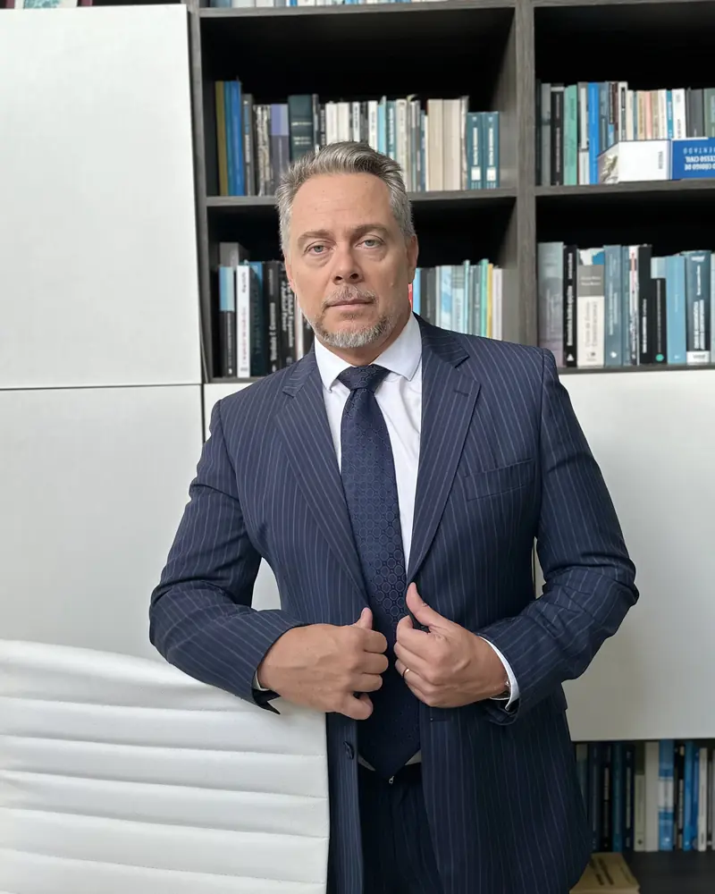 Leandro Corrêa de Oliveira, de terno, apoiado em uma cadeira diante de uma estante de livros